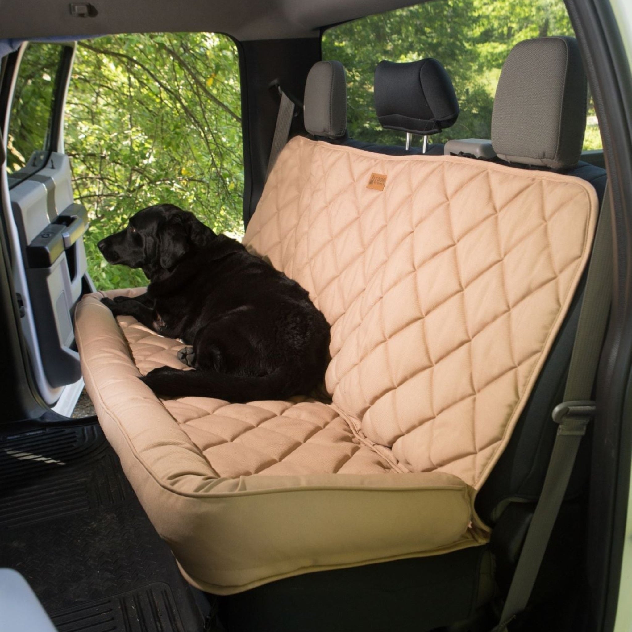 A black dog laying on a tan Farm to Pet Accessory Crew Cab Truck back seat protector in a vehicle with greenery outside.