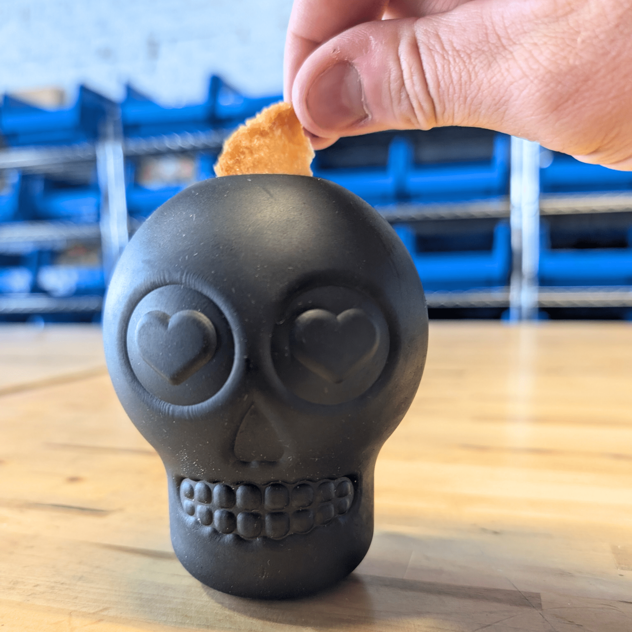 Farm to Pet Dog Treat Dispenser Black skull-shaped with heart eyes on a wooden surface a hand putting in a chip.