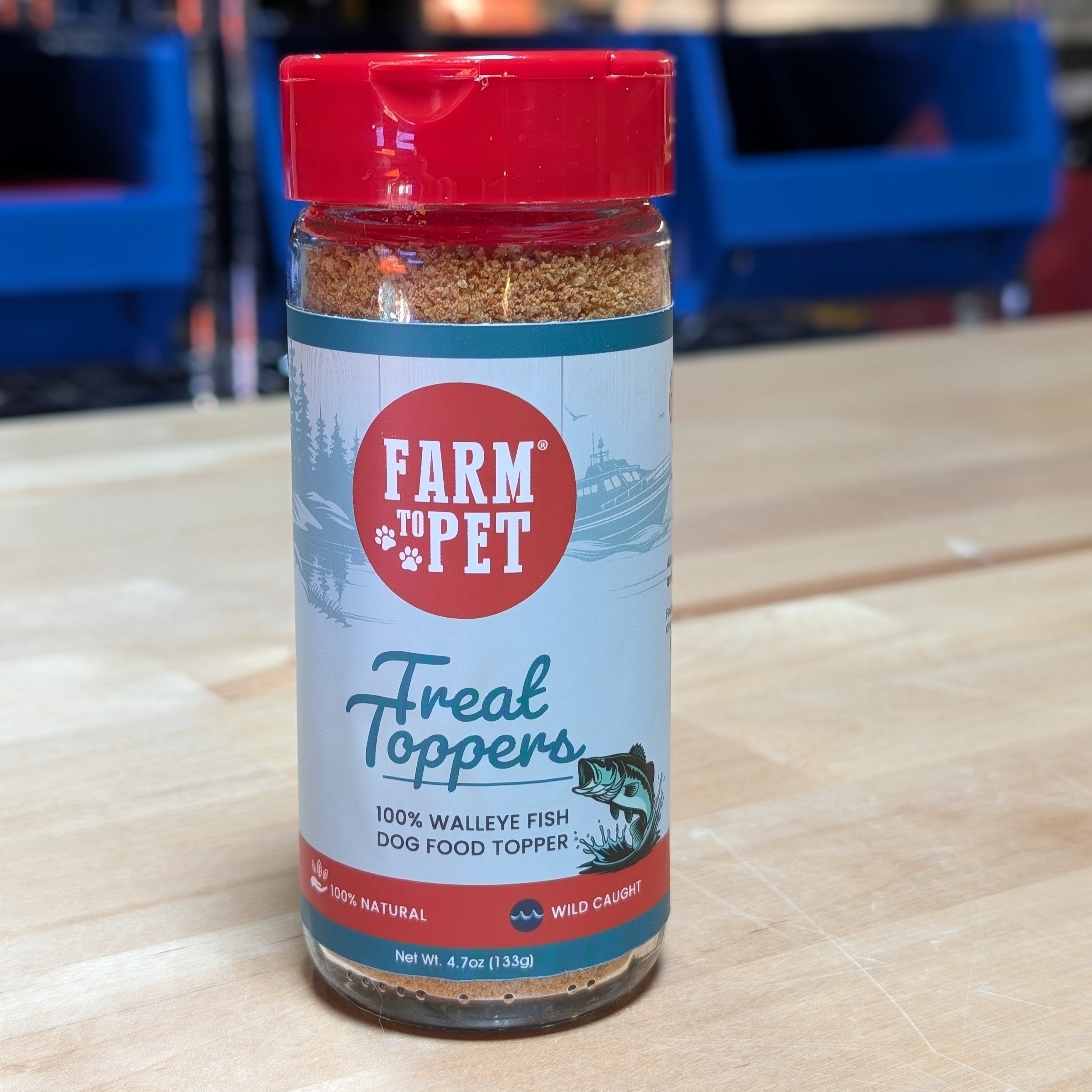 A jar of Farm to Pet Fish Treat Toppers on a wooden surface with blue storage bins in the background.