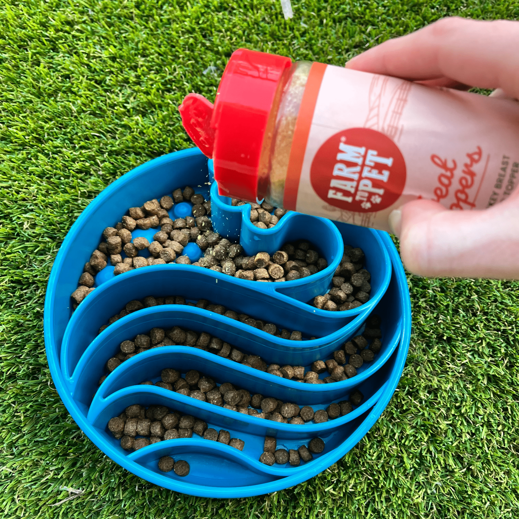 Hand pouring a product from Farm to Pet into a blue pet feeder filled with kibble on grass.