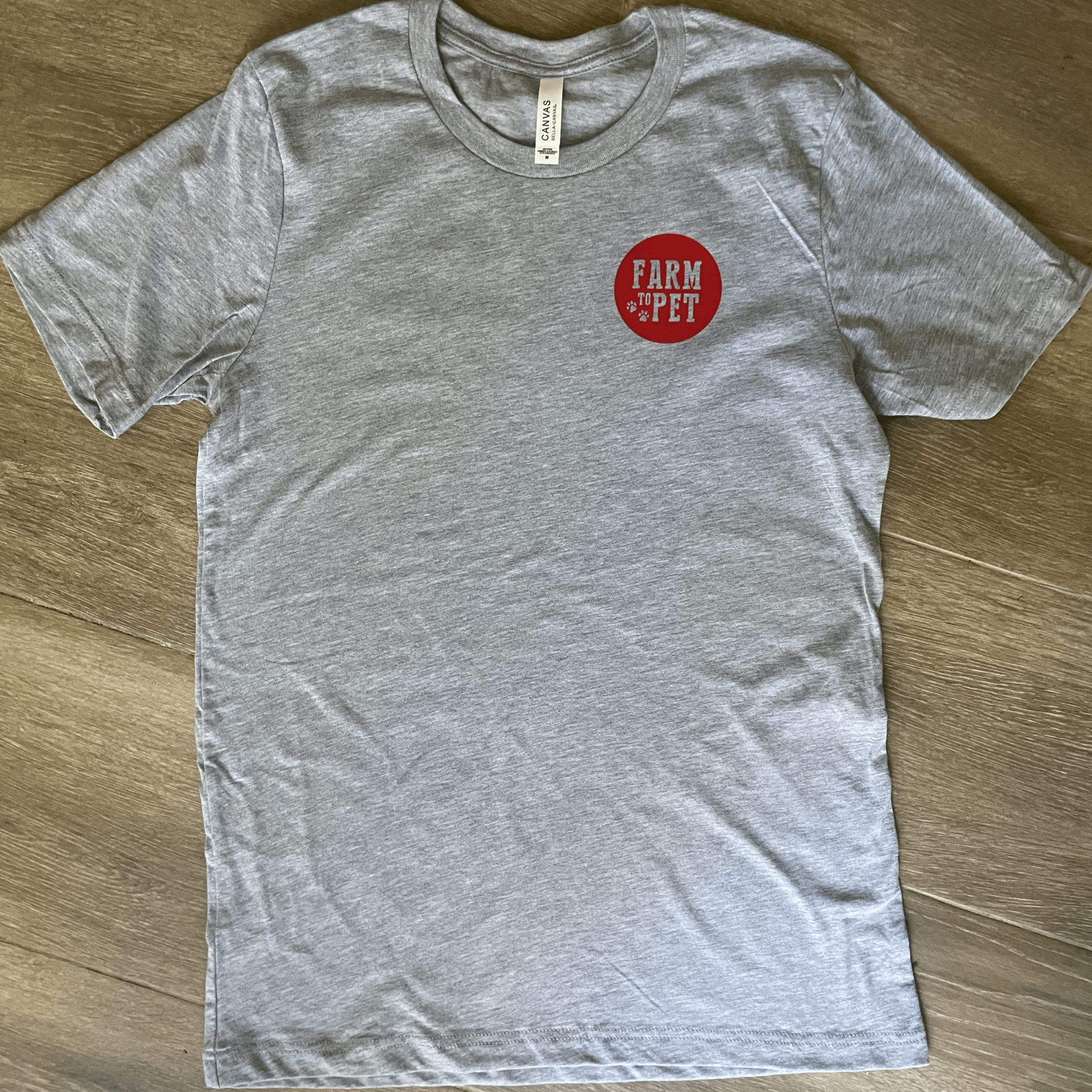 The front of a gray T-shirt with a red Farm to Pet logo laying on a wooden surface.
