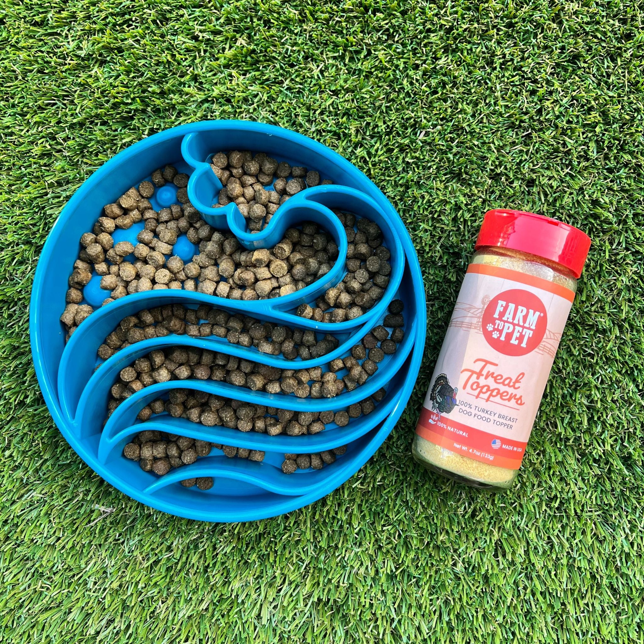 A Farm to Pet Blue Wave pet food bowl with kibble and a jar of Farm & Pet Turkey Treat Toppers on grass.
