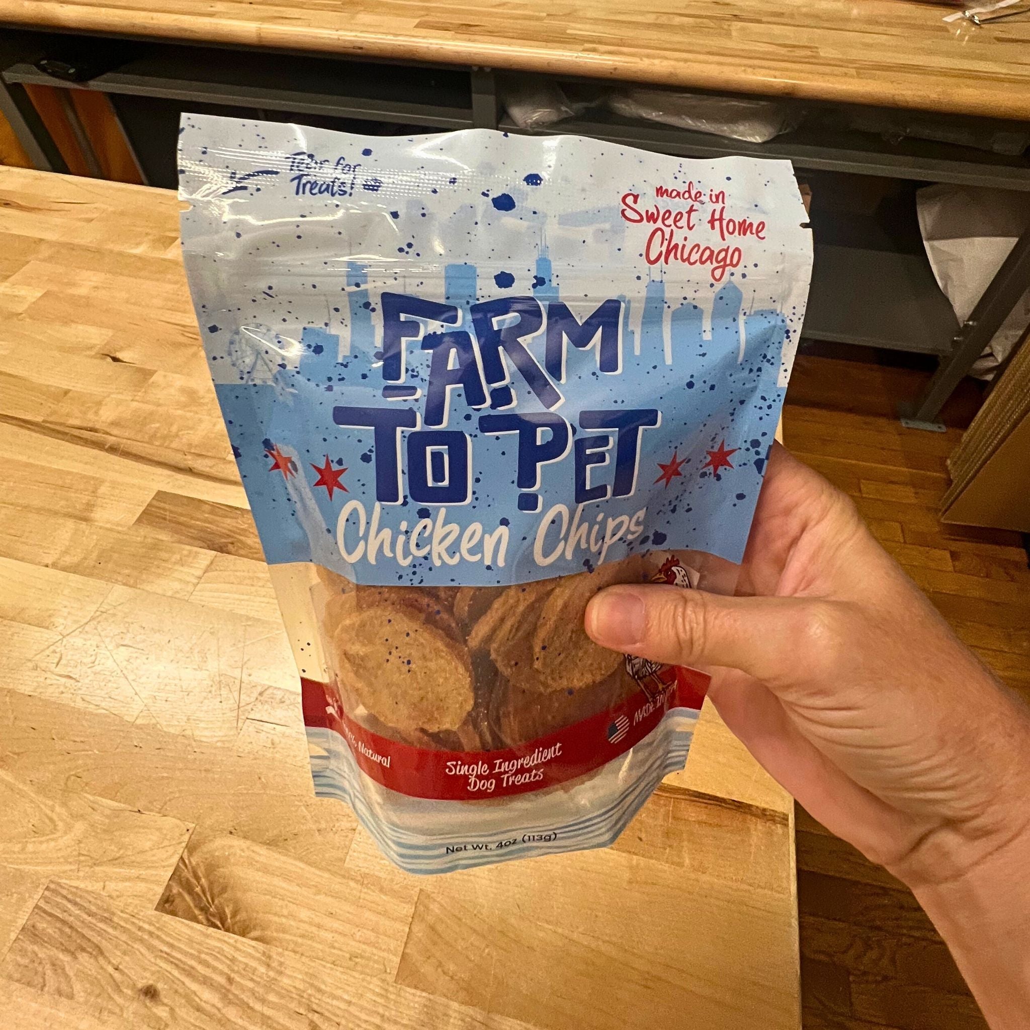 A product picture of Farm to Pet Chicago Chicken Chips Bag held by a hand above a wooden table.
