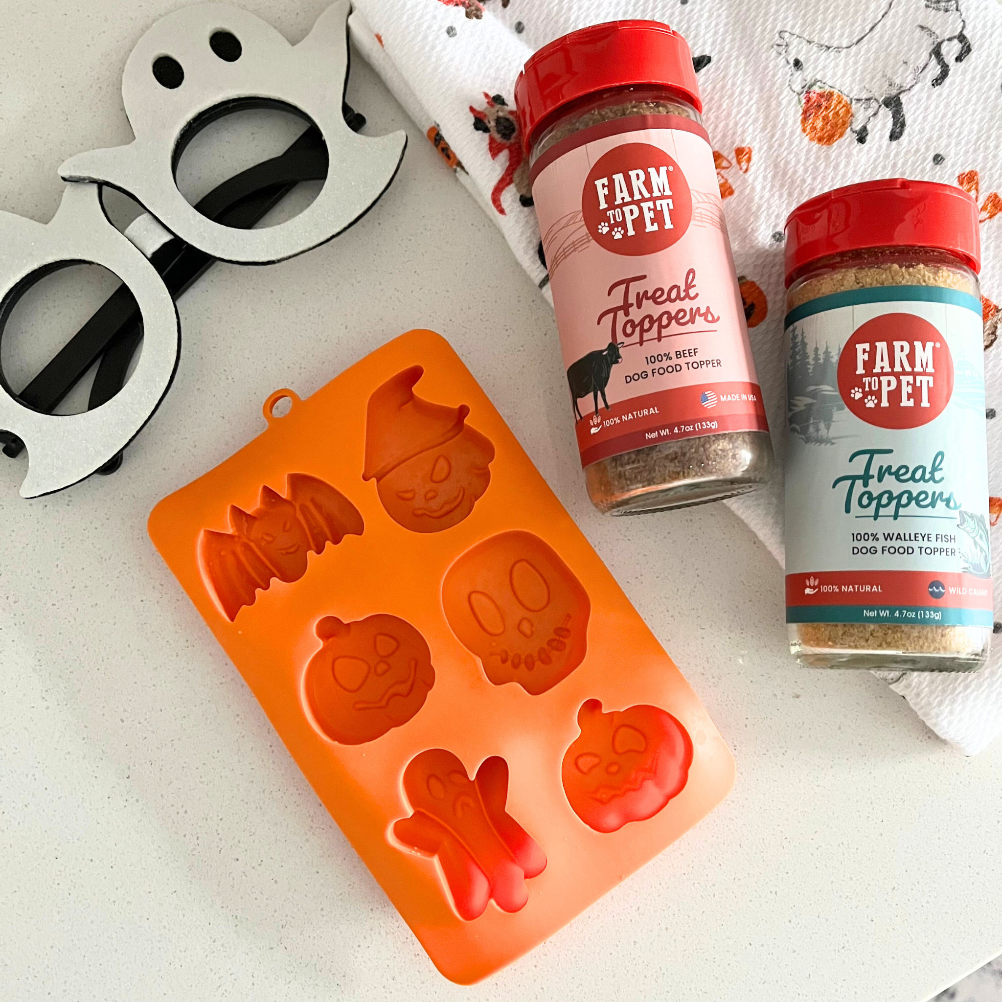 Two jars of 'Farm to Pet' treat toppers beef and fish mix and a orange silicone mold with Halloween designs on a white surface.