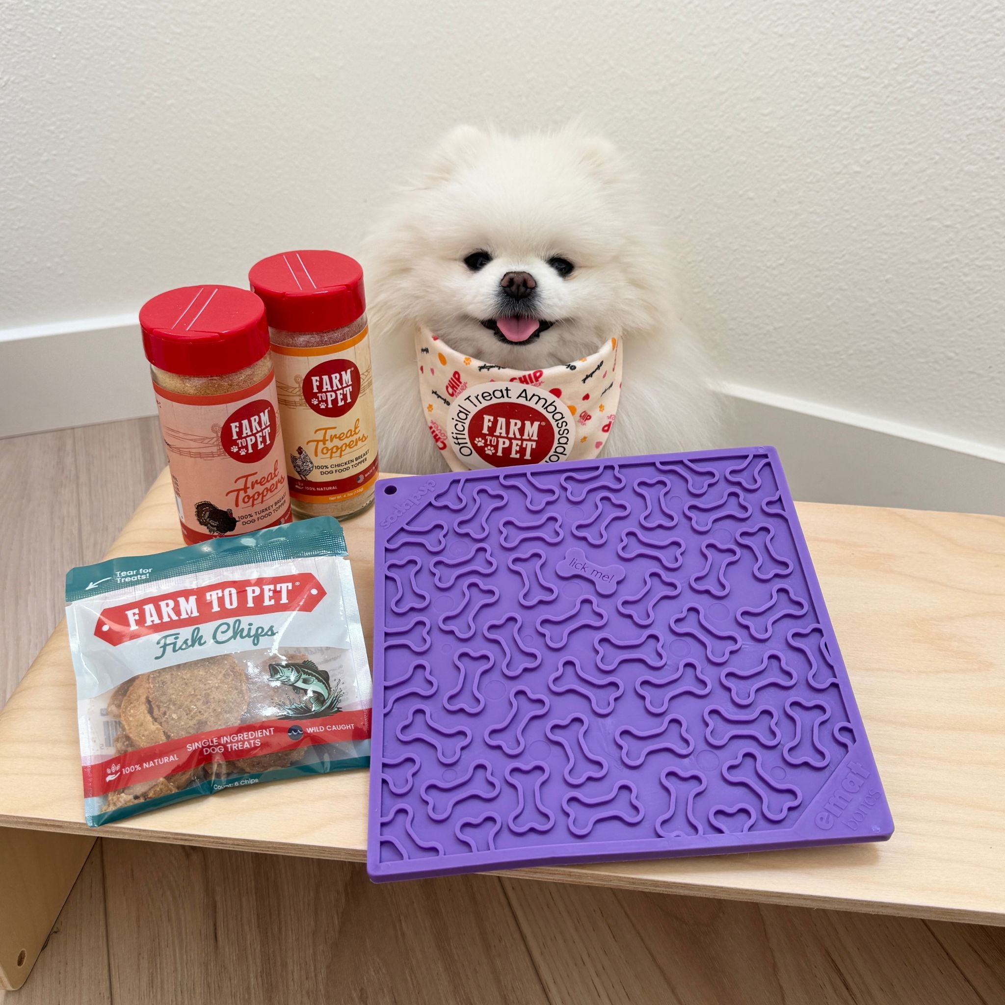 Farm to Pet purple dog bone lick mat with a white dog wearing a Farm to Pet bandana and Farm to Pet Fish Chips and Treat Toppers on a table.