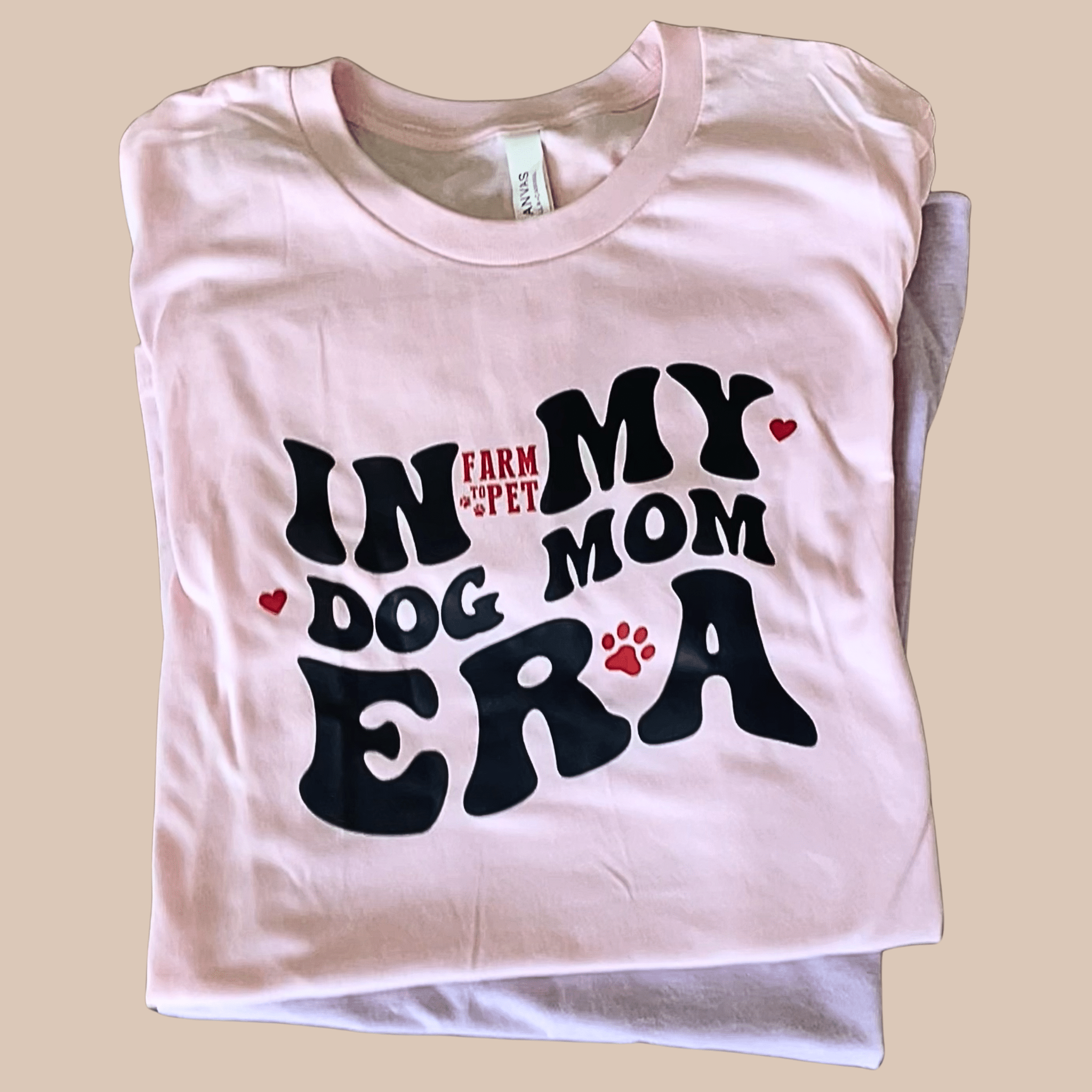 A folded Farm to Pet T-shirt with 'In My Dog Mom Era' text on a beige background.
