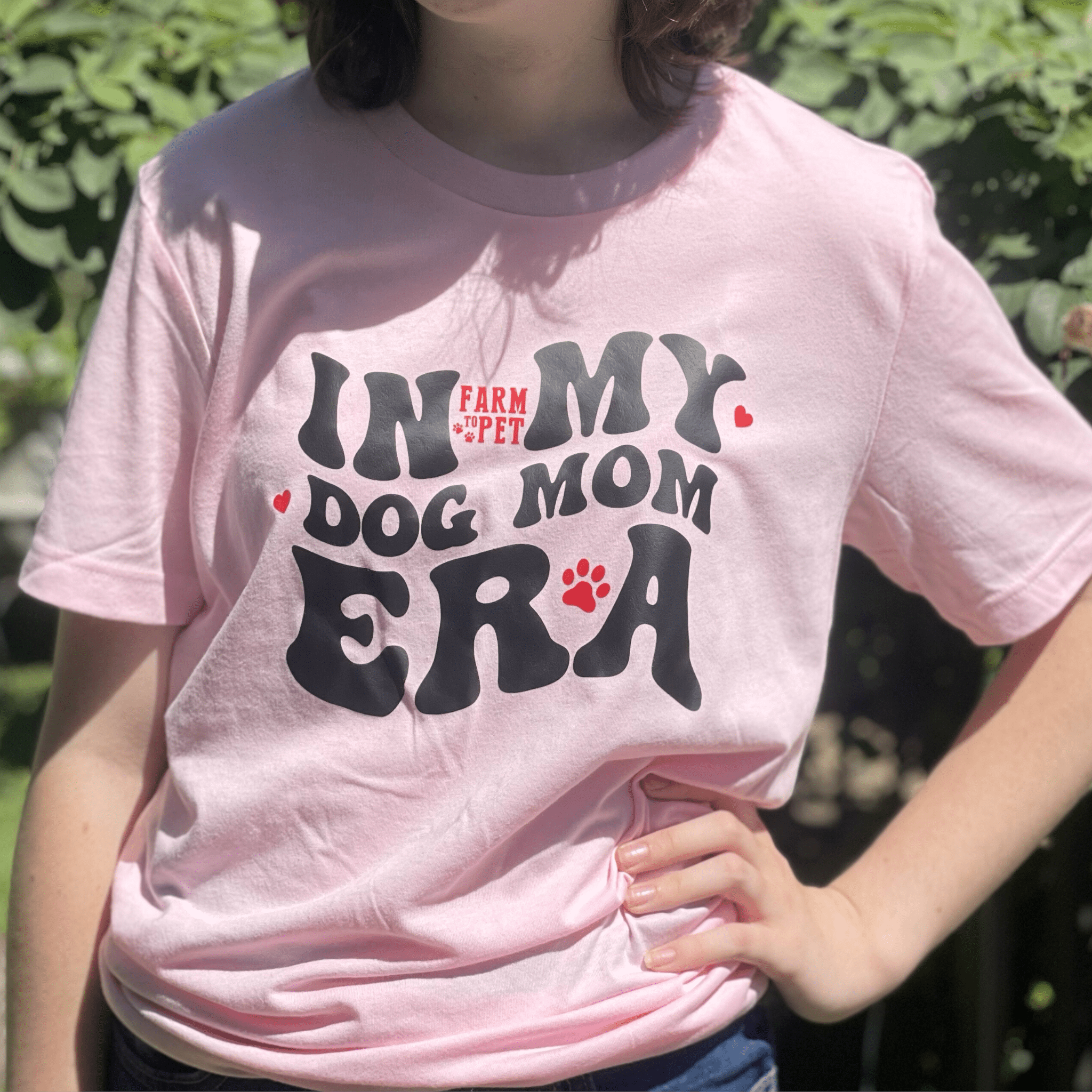 A woman wearing a Farm to Pet pink t-shirt with 'In My Dog Mom Era' text, standing outdoors.