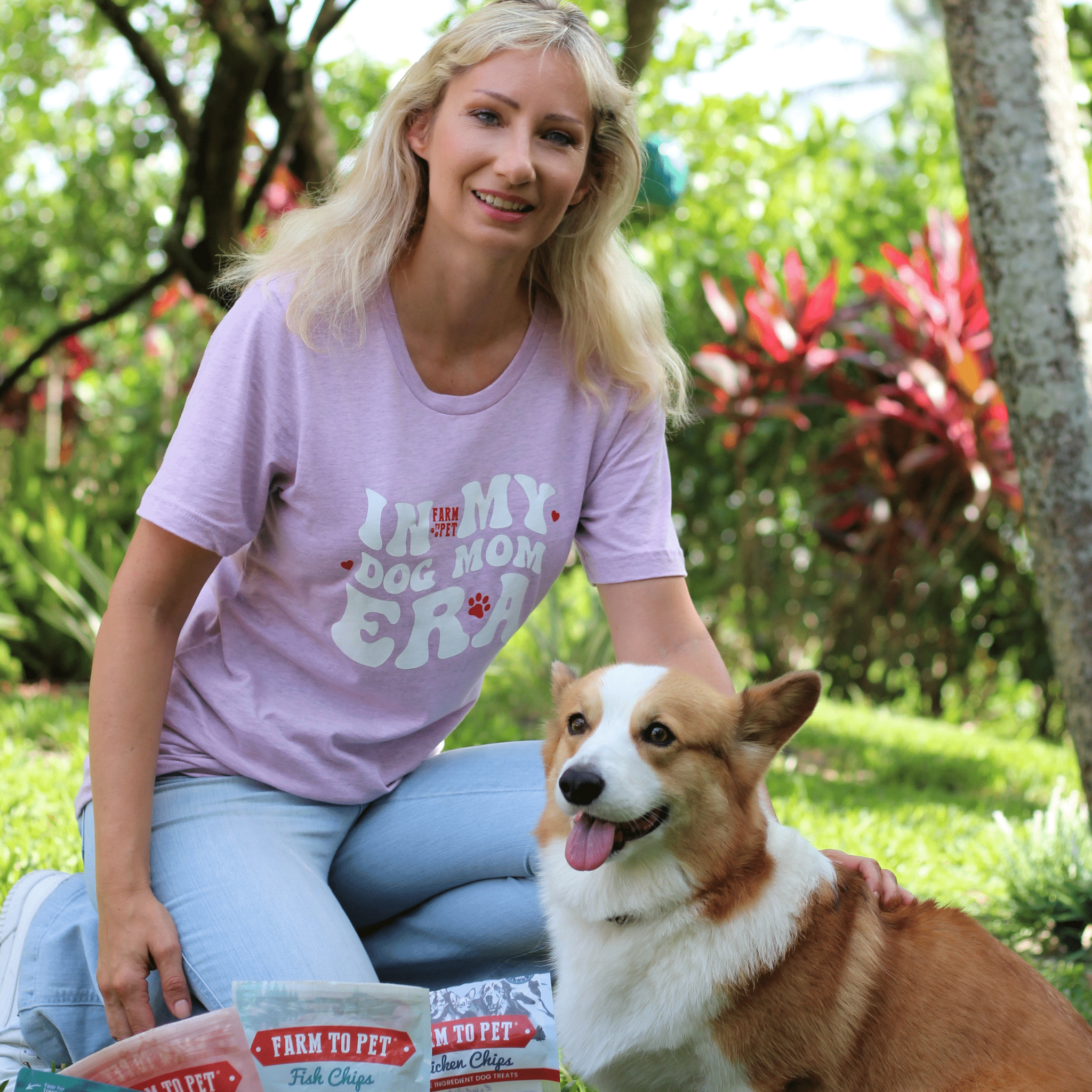 A woman wearing a purple Farm to Pet  t-shirt with a dog-themed design, sitting outdoors with a corgi.
