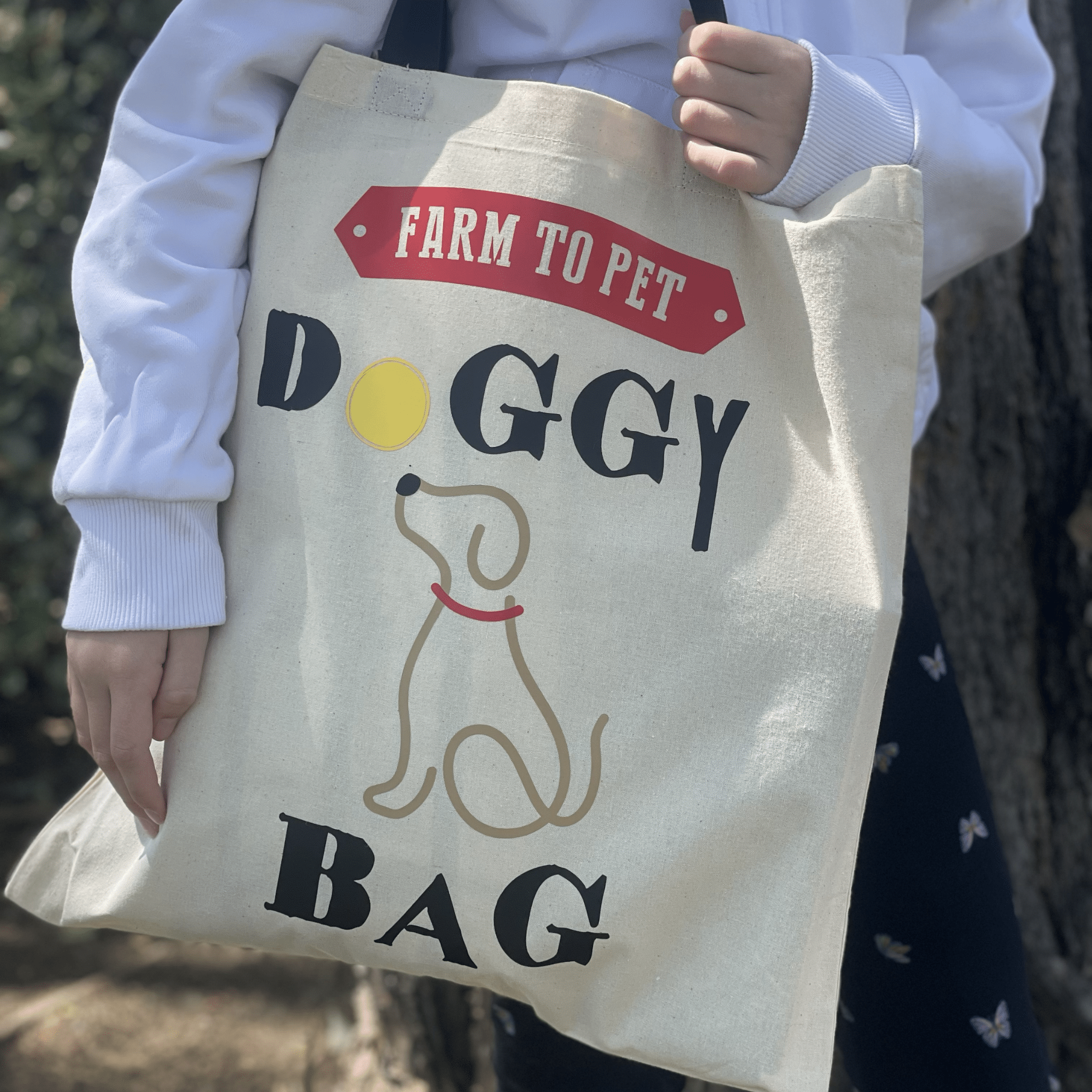 A white and black tote bag with Farm to Pet Doggy Bag text and design held by a person outside.