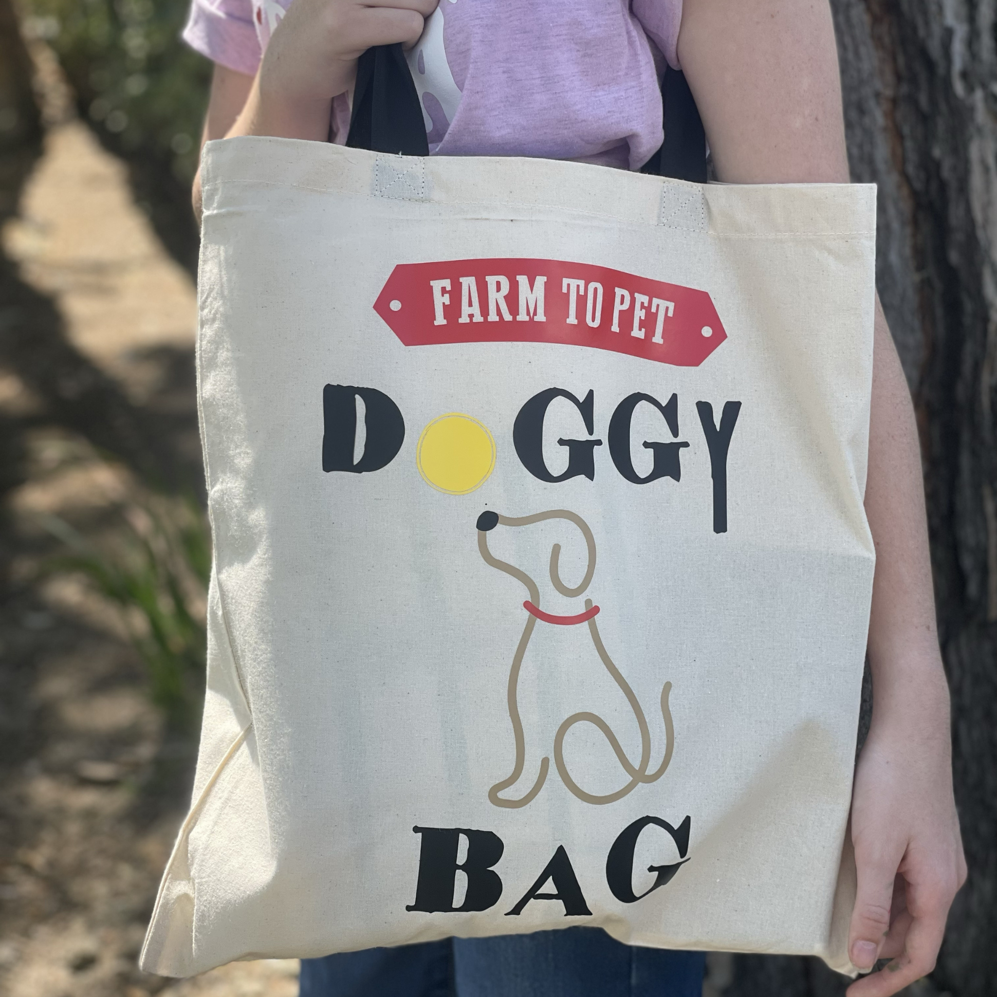 A person holding a Farm to Pet Doggy Tote Bag leaning against a tree.