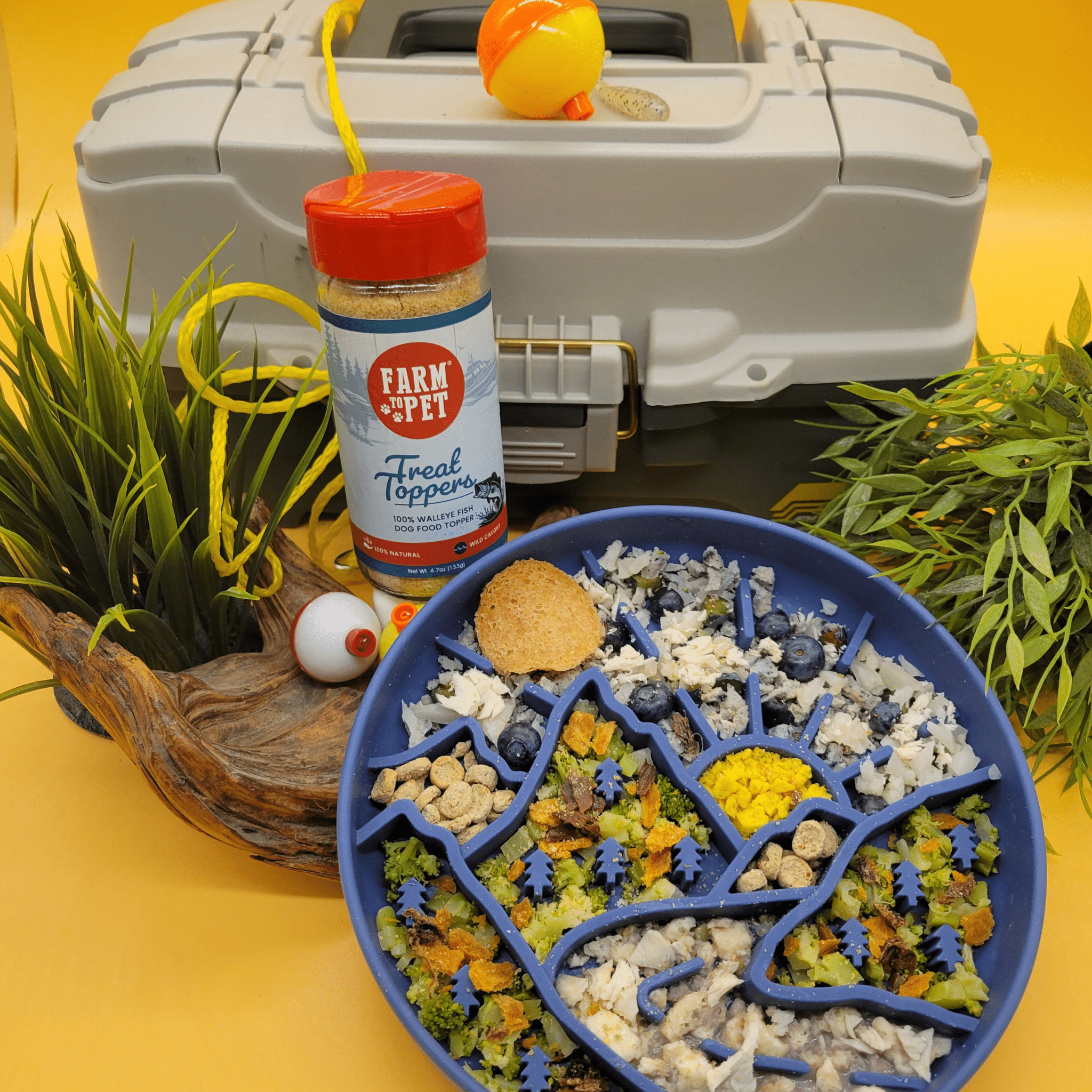 Farm to Pet Fish Topper sprinkled on food with colorful toppings on a blue plate, next to a gray fishing tacklebox on a yellow background.