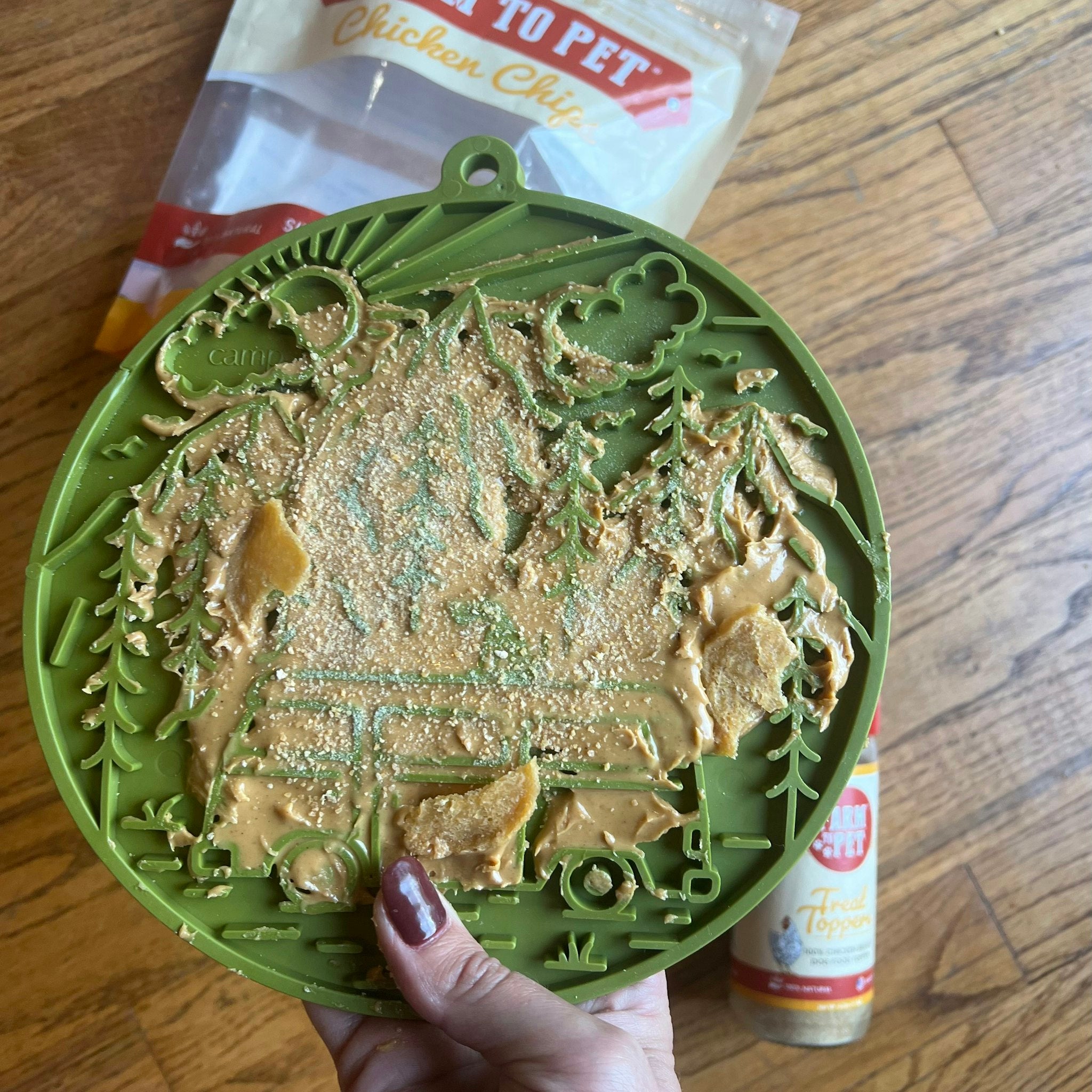 Farm to Pet green camp Lick Mat held by a hand filled with chicken chips and sprinkled with chicken treat toppers on a wooden background.