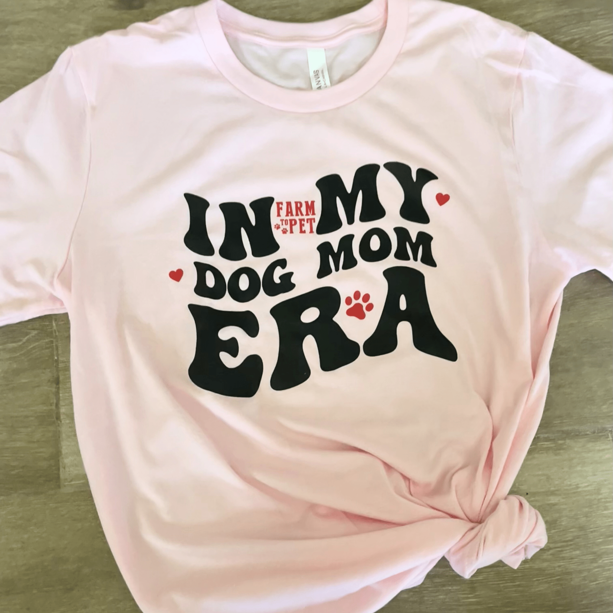 A pink Farm to Pet In MY Dog Mom ERA T-shirt laying on a wooden suface.