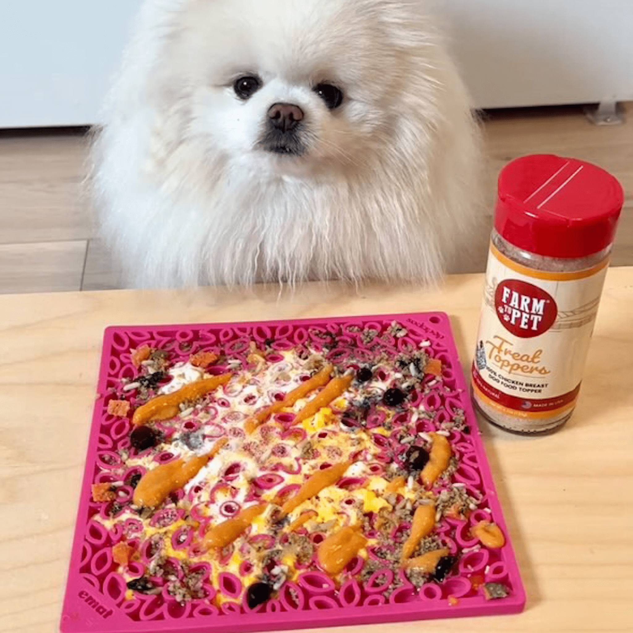A White dog looking at a Farm to Pet  pink lick mat with chicken treat topper jar product on a wooden table.
