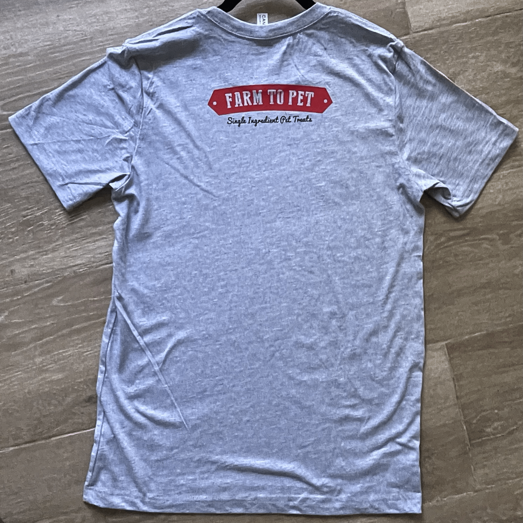 The Back of a gray T-shirt with Farm to Pet red logo laying on a wooden surface