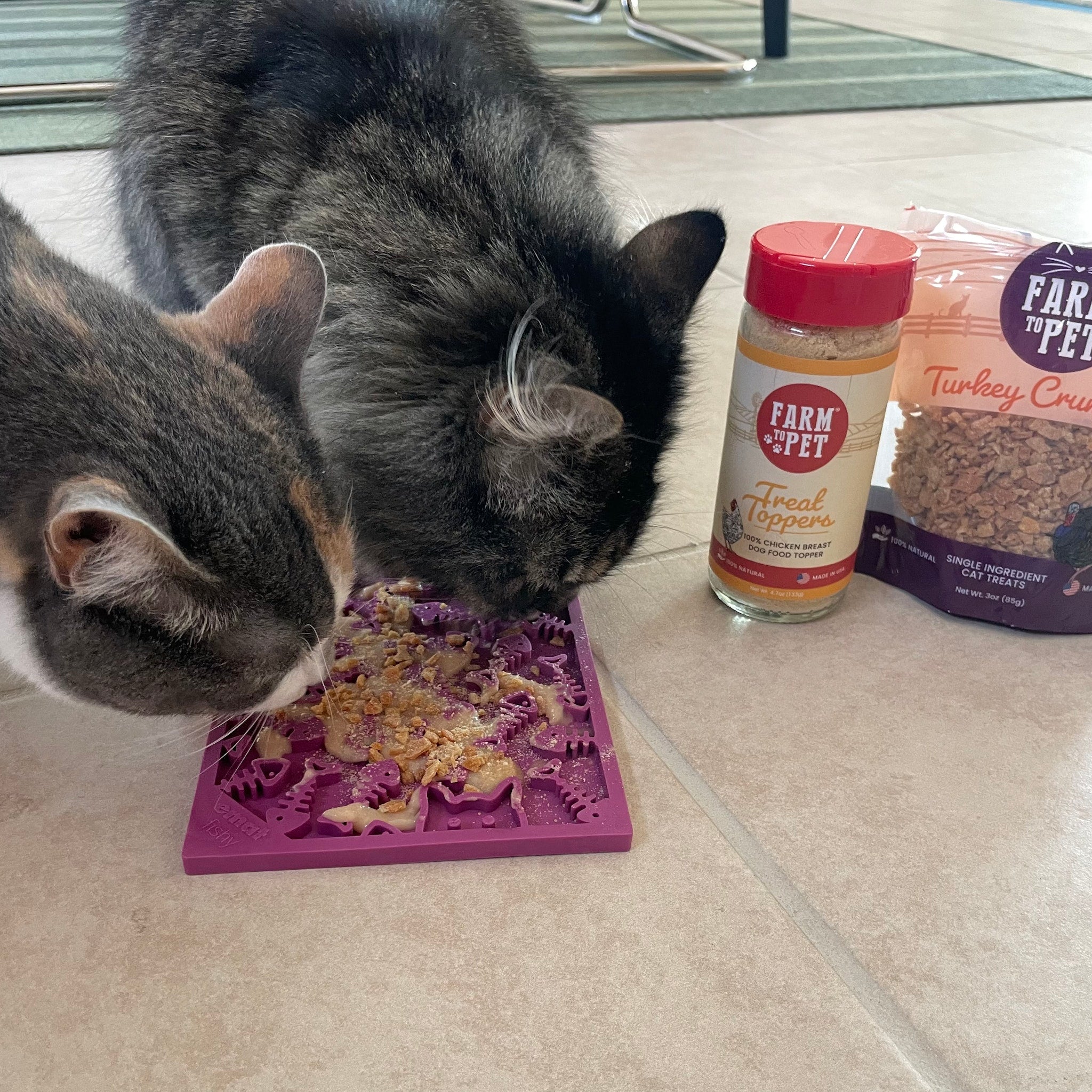Two cats eating from a Farm to Pet maroon lick mat with a jar of Farm to Pet treat topper and a bag of turkey crumbles in the background.