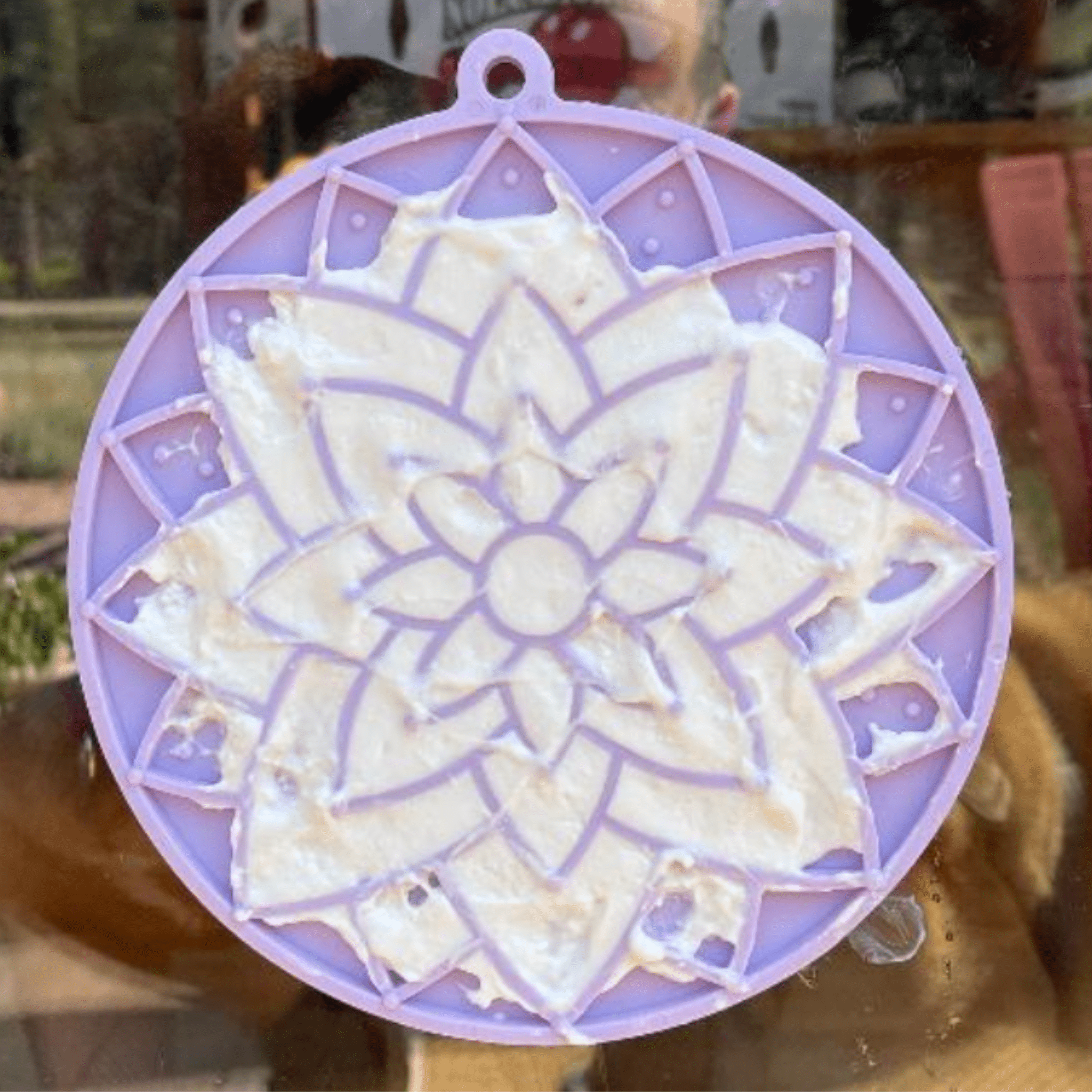 A Farm to Pet Purple Lotus Dog Lick Mat with a floral design filled with yogurt and stuck to a window.