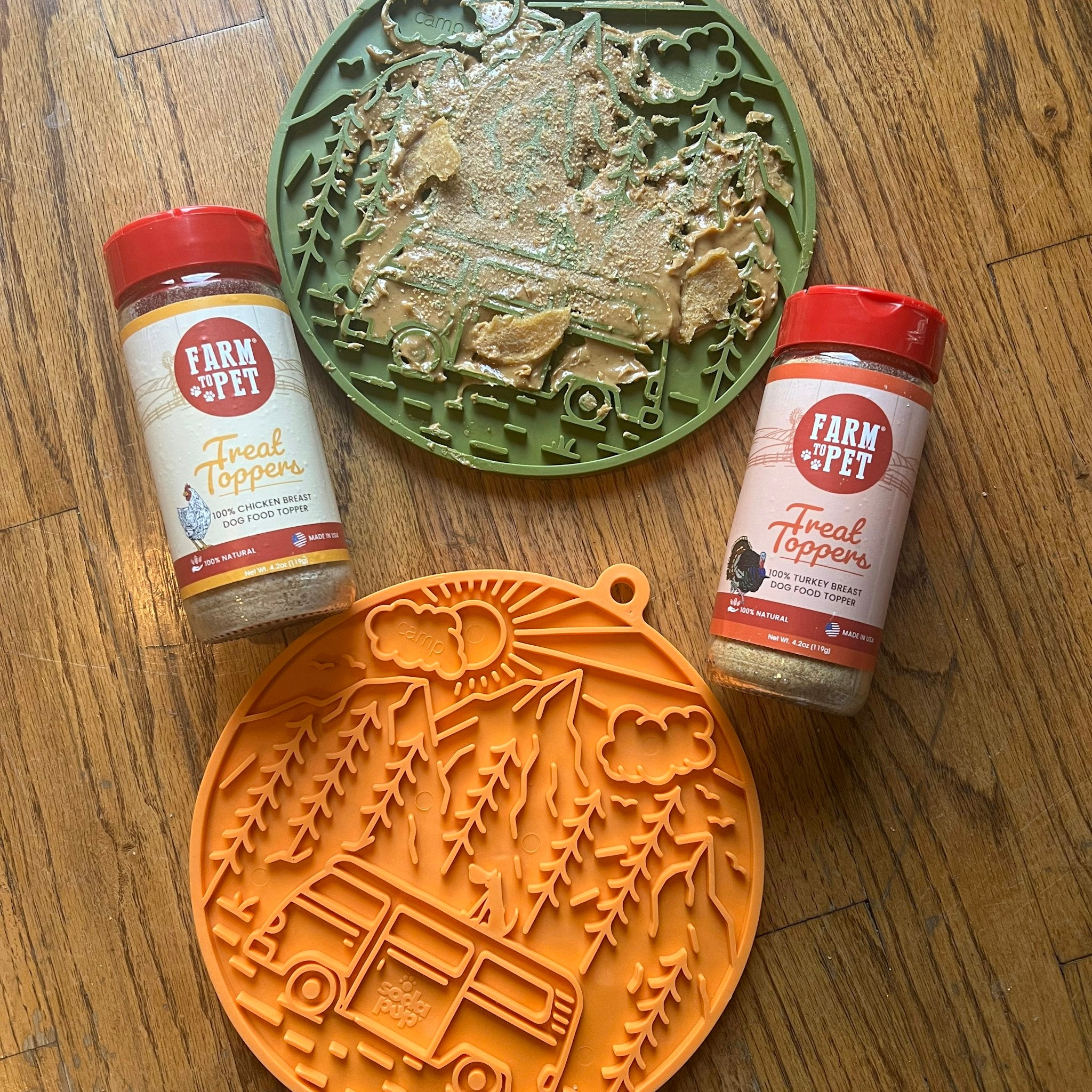 Two Farm to Pet Dog Lick Mats green (filled with treats) and orange camp design with Farm to Pet Chicken and Turkey Treat Toppers on a wooden table.