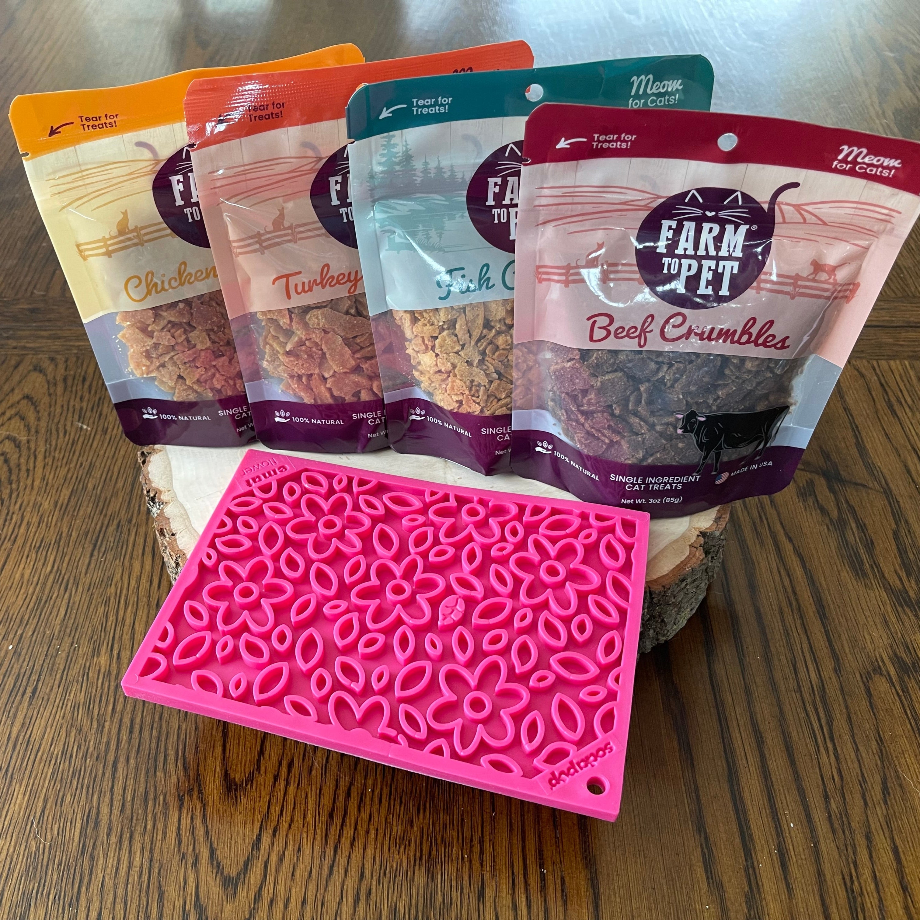 Four packages of Farm Pet Cat Crumbles Chicken Turkey, Fish and Beef ,  treats on a wooden surface with a Farm to Pet  Pink Flower Power Lick Mat front of bags. 
