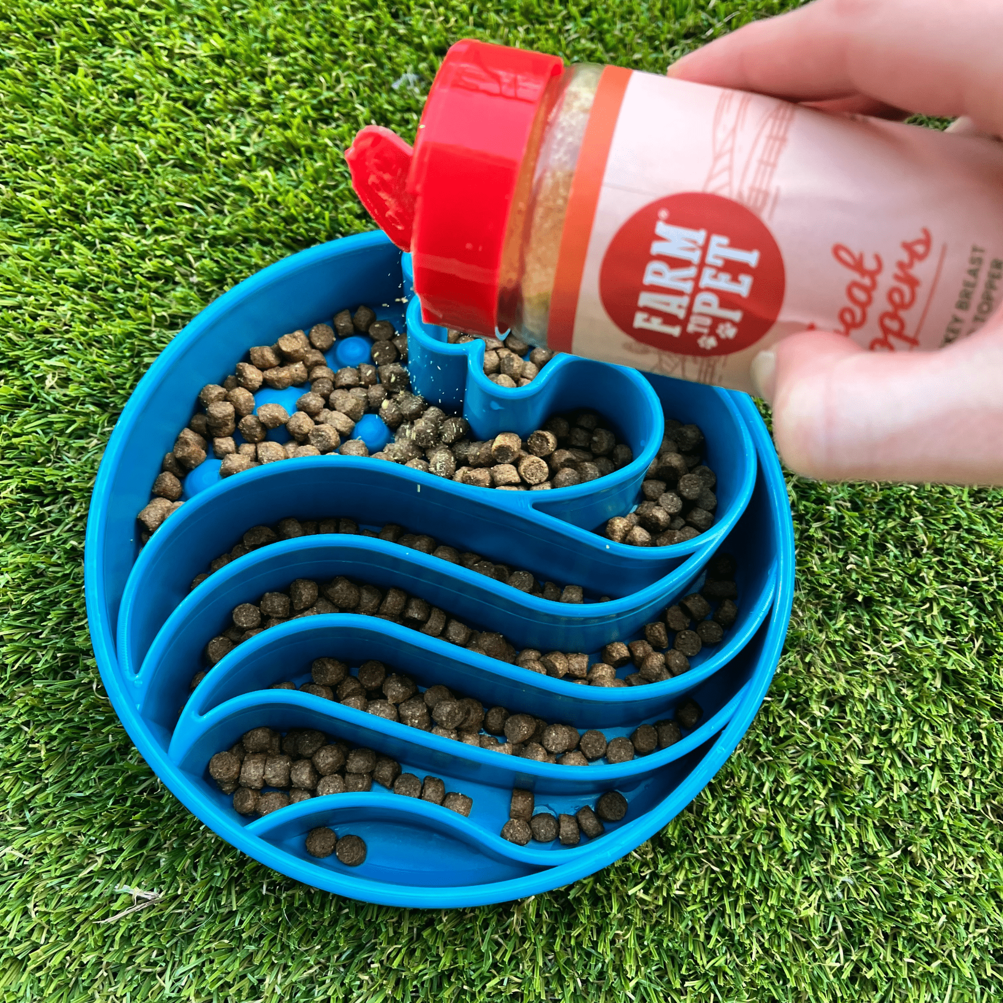 Farm to Pet Slow Feeder Dog Bowl Blue Wave with food being  sprinkled with Farm to Pet treat topper from a jar on a grass background.