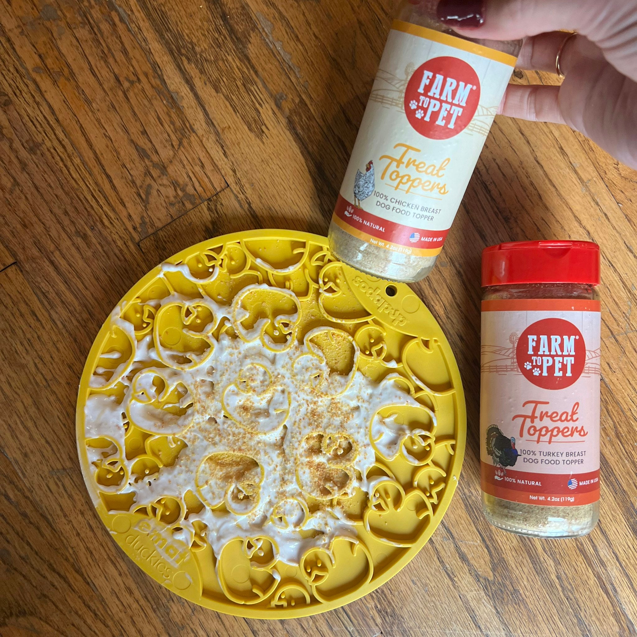A Farm to Pet yellow duckies lick mat spread with yogurt and sprinkle with treat toppers laying on a wood surface.