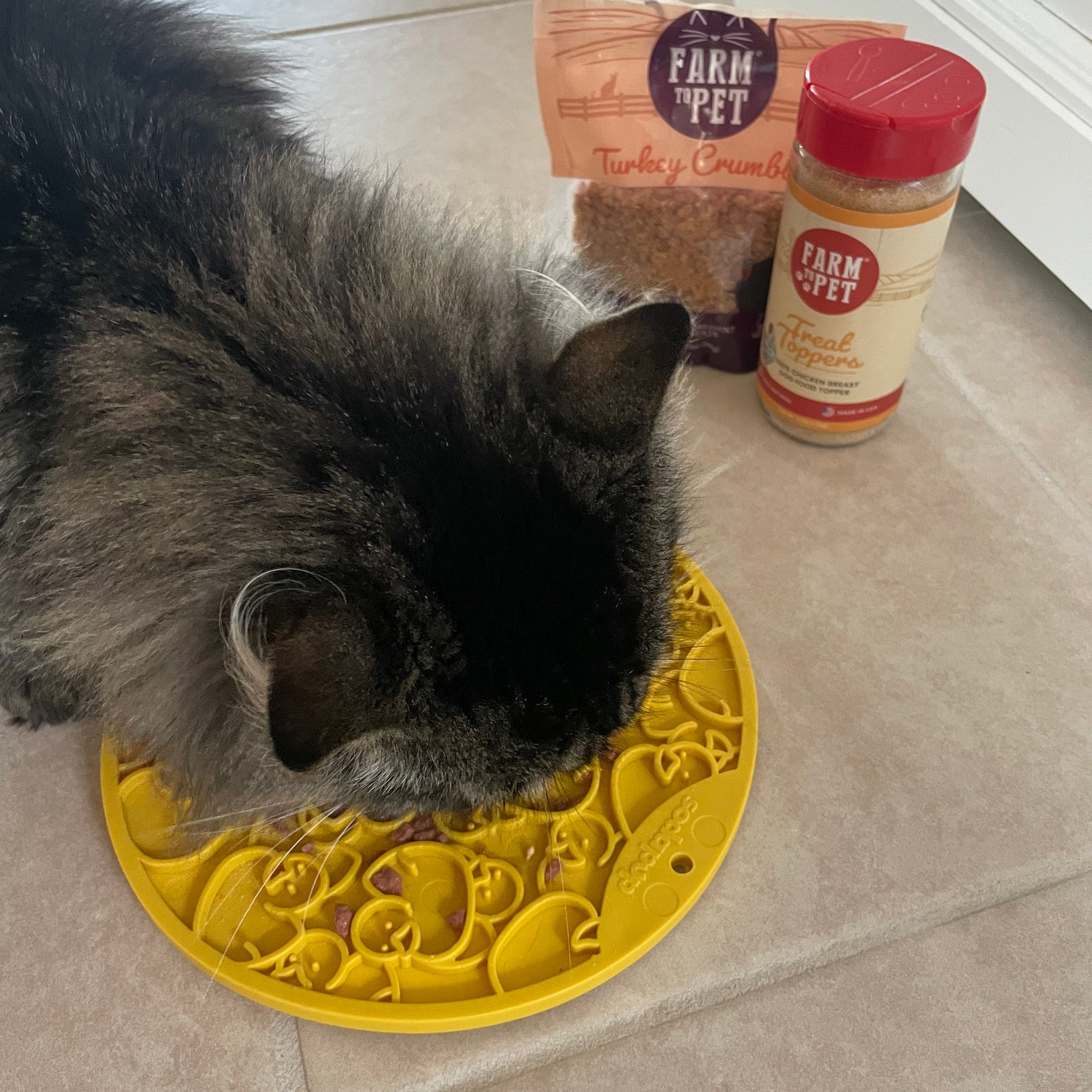 A gray cat licking a Farm to Pet  yellow duckies lick mat with 'Farm to Pet' products in the background.