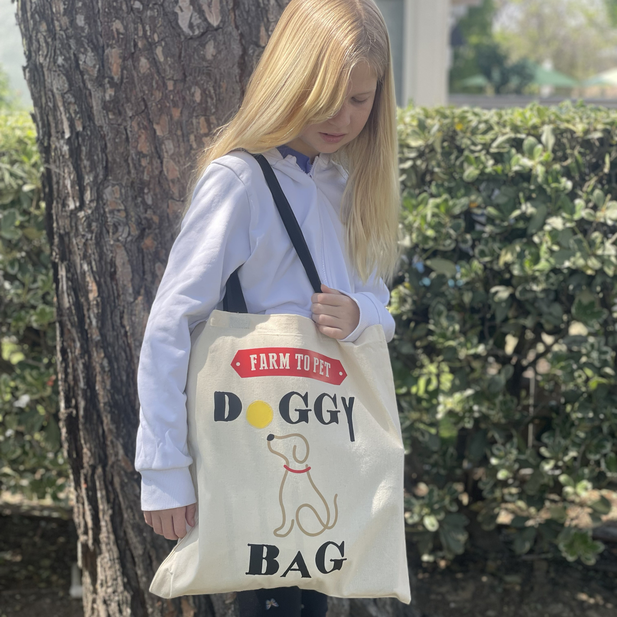 girl holding a tote bag with 'Farm to Pet Doggy Bag' text and dog illustration outdoors.