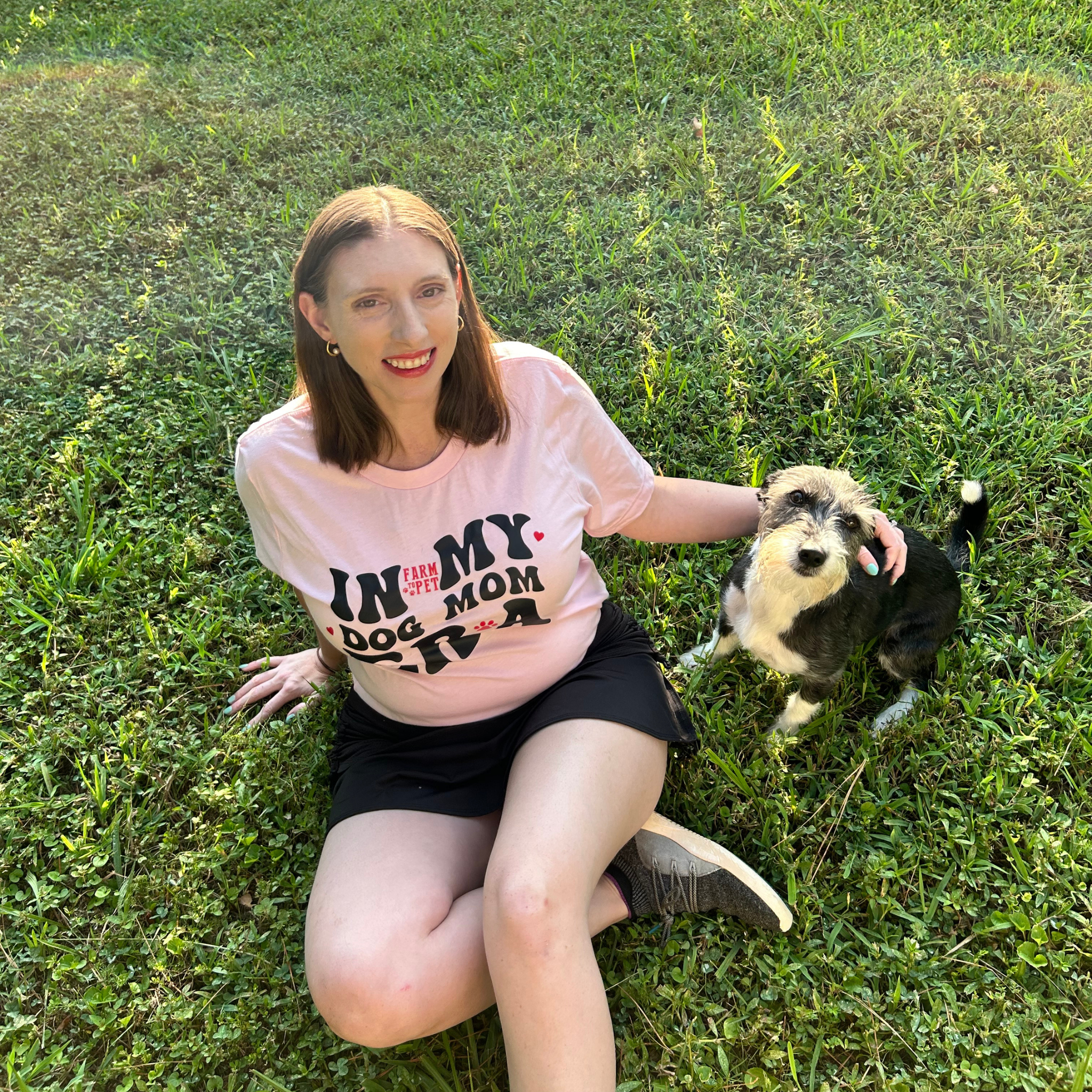 Woman sitting on grass with a small dog, wearing a pink t-shirt with text.