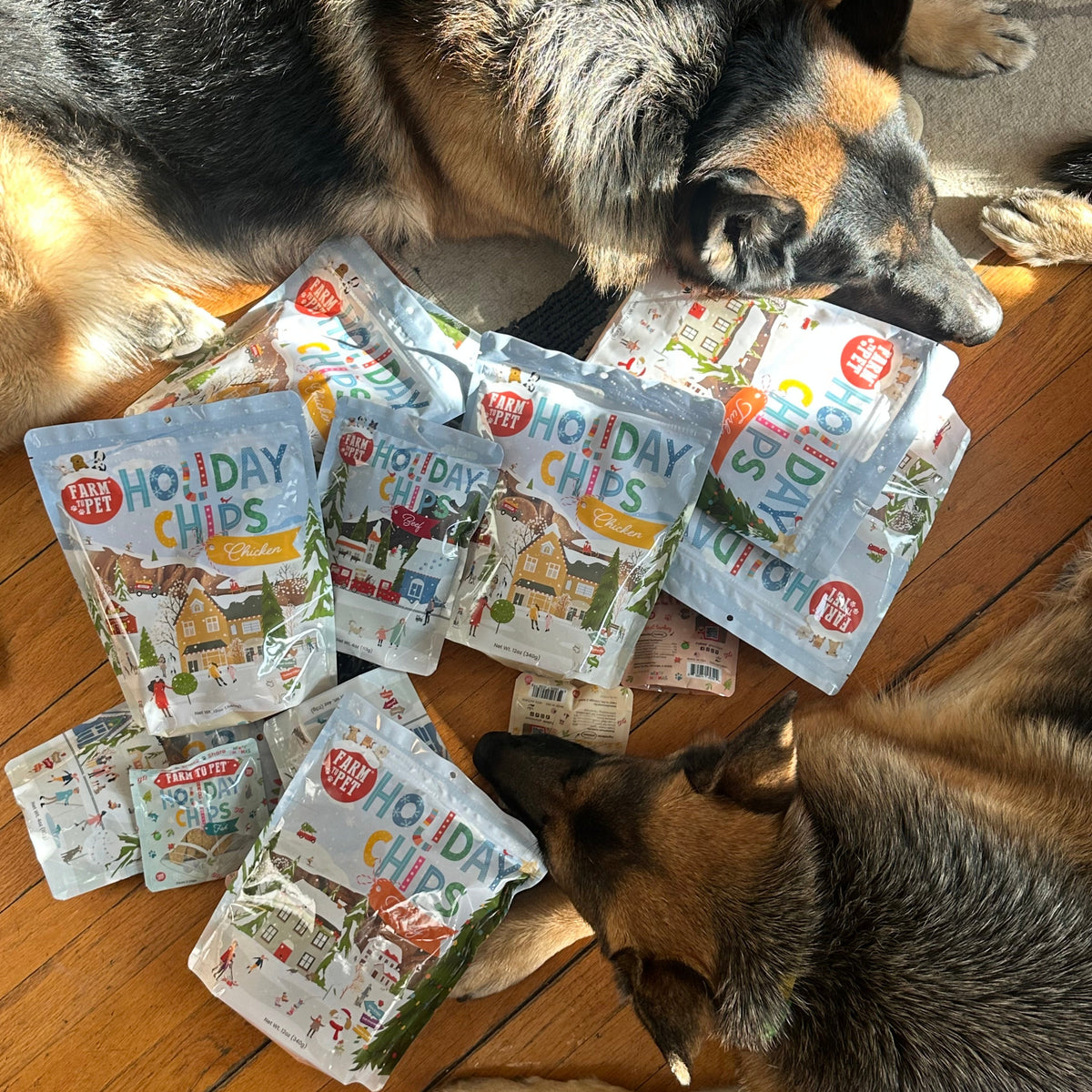Two dogs next to a pile of Farm to Pet Holiday Chips bags, healthy Christmas dog treats