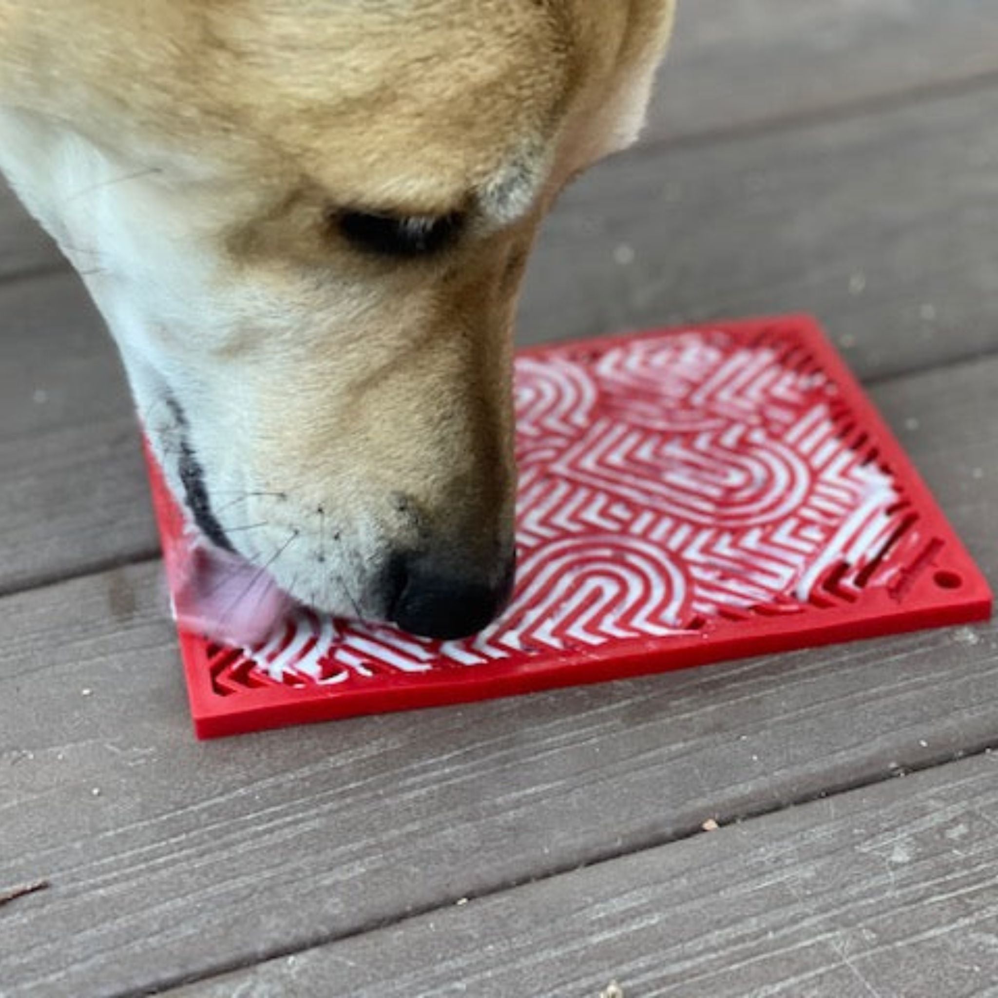 A dog licking a Farm to Pet red hearts lick mat  on a wooden deck.